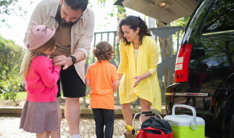 Uma família se organiza perto de um carro preto, preparando-se para um passeio.