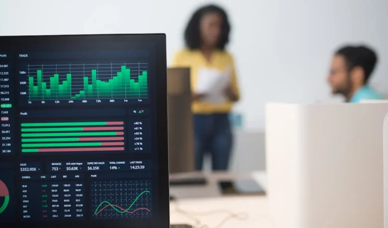 Tela de computador exibindo gráficos financeiros e duas pessoas em um escritório.