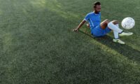 Homem sentado em campo de grama sintética, em traje de futebol azul, controlando uma bola.
