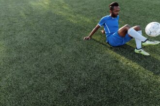 Homem sentado em campo de grama sintética, em traje de futebol azul, controlando uma bola.
