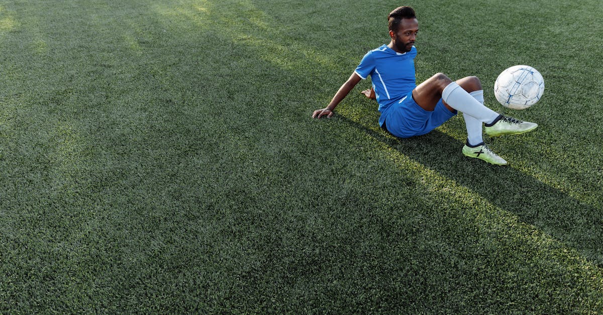Homem sentado em campo de grama sintética, em traje de futebol azul, controlando uma bola.
